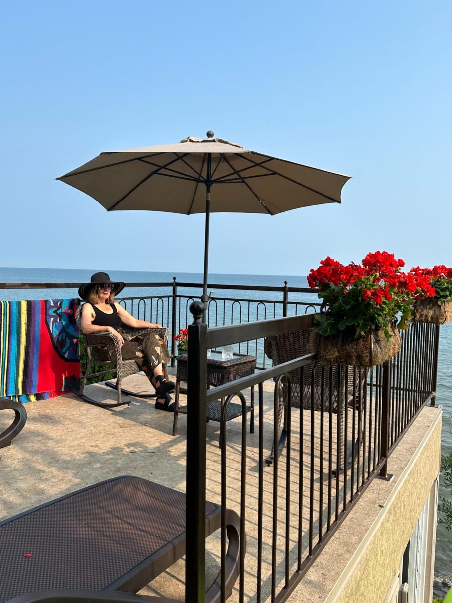 Mole End Upper Patio Seating and Lake Views Lakeside patio with lounge chairs, umbrella, bright flowers, and panoramic views of Lake Ontario at Wind in the Willows Oasis.